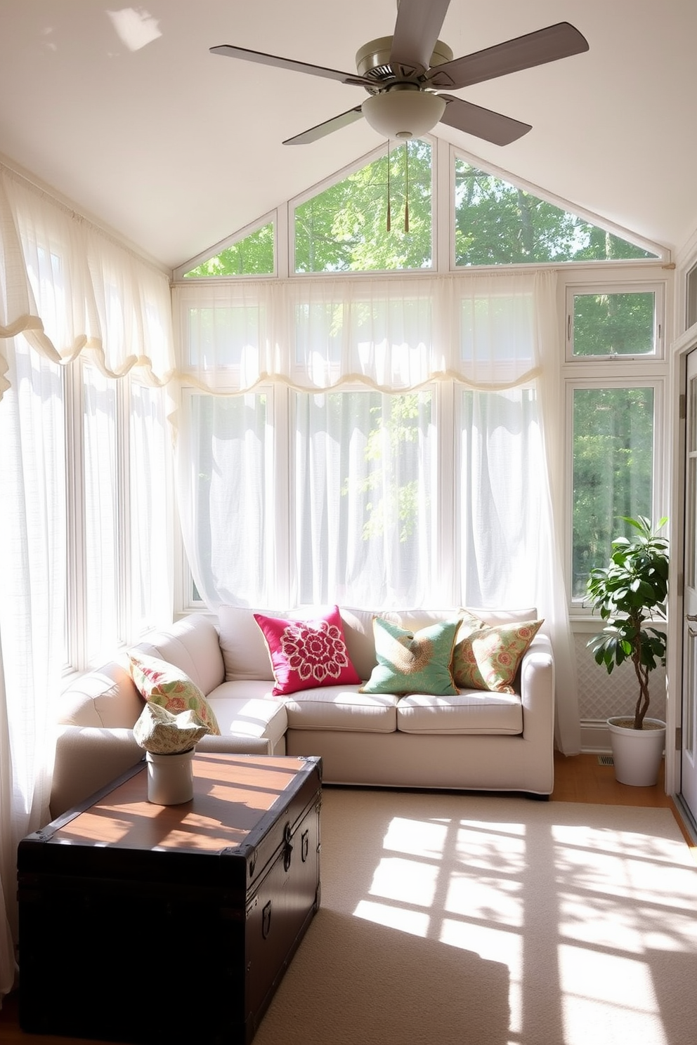 A bright and airy sunroom filled with natural light. The space features large windows adorned with sheer white curtains, allowing the summer sun to filter through. In the center, a comfortable seating area is arranged with a plush, light-colored sofa and vibrant throw pillows. A vintage trunk sits in the corner, serving as unique storage and adding character to the room.