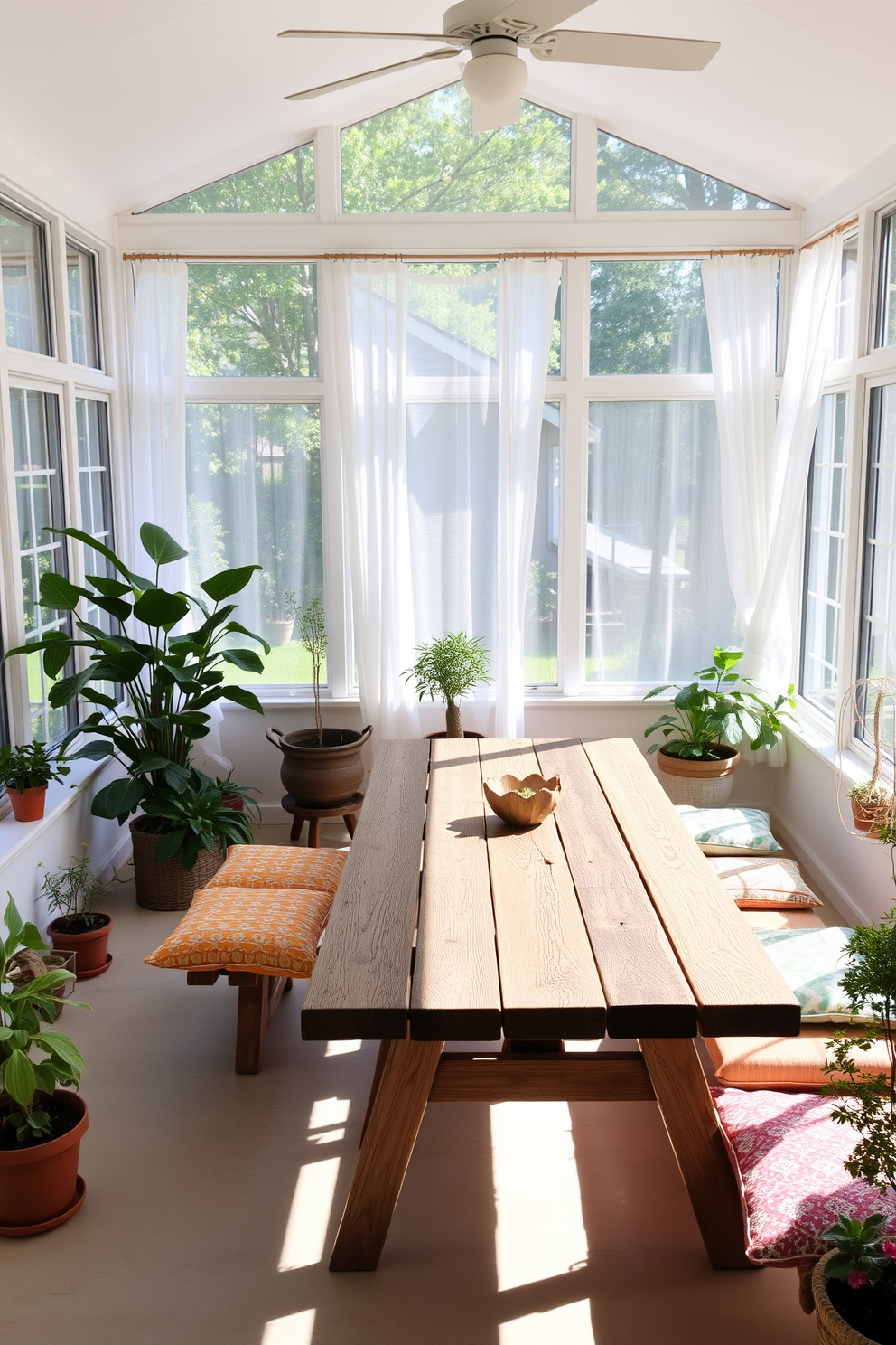 A bright summer sunroom filled with natural light. A large picnic table made of weathered wood serves as the centerpiece, surrounded by colorful cushions for casual dining. Large windows adorned with sheer white curtains allow the sun to filter in gently. Potted plants in various sizes are placed around the room, adding a touch of greenery and freshness.